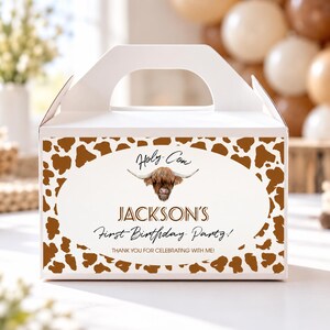 May include: A white cardboard gable box featuring a brown cow print border. The center oval graphic displays a Highland cow illustration with the text Holy Cow, JACKSON'S First Birthday Party!, and THANK YOU FOR CELEBRATING WITH ME!