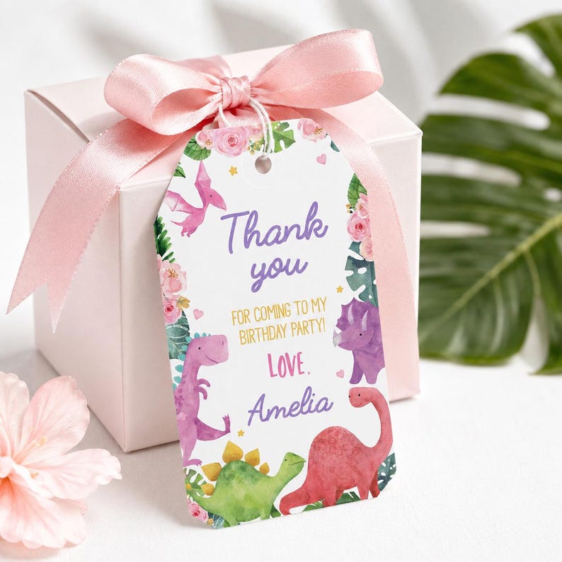K&ouml;nnte beinhalten: Eine wei&szlig;e Geschenkbox mit einem hellrosa Satinband und einem rechteckigen Papieranh&auml;nger, der mit Aquarellillustrationen von Dinosauriern, rosa Blumen und gr&uuml;nen Bl&auml;ttern verziert ist. Der Text lautet Thank you FOR COMING TO MY BIRTHDAY PARTY! Love. Amelia.