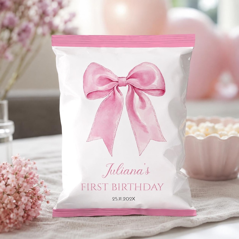 May include: A white snack bag with pink top and bottom edges, featuring a large pink watercolor bow illustration in the center. The text reads Juliana's FIRST BIRTHDAY and 25.11.202X in a pink serif font. The bag has a smooth, sealed plastic finish.