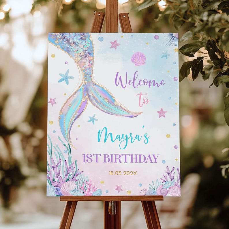 May include: A welcome sign for a 1st birthday party with a mermaid tail design. The sign is decorated with glitter, seashells, and stars. The text on the sign reads "Welcome to Mayra's 1st Birthday 18.03.202X".