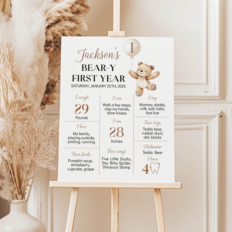 May include: A white milestone birthday sign titled Jackson's BEAR-Y FIRST YEAR with a brown teddy bear illustration, a balloon with the number 1, and text detailing weight in pounds, height in inches, favorite foods, songs, toys, and milestones.