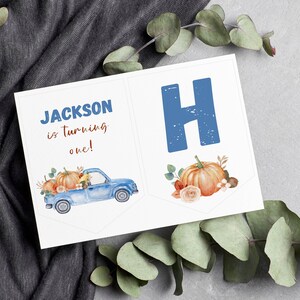 May include: A white rectangular banner featuring a blue truck carrying orange pumpkins and flowers, with the text "JACKSON is turning one!" in blue and brown, alongside a second banner section displaying a large blue letter H and an orange pumpkin.