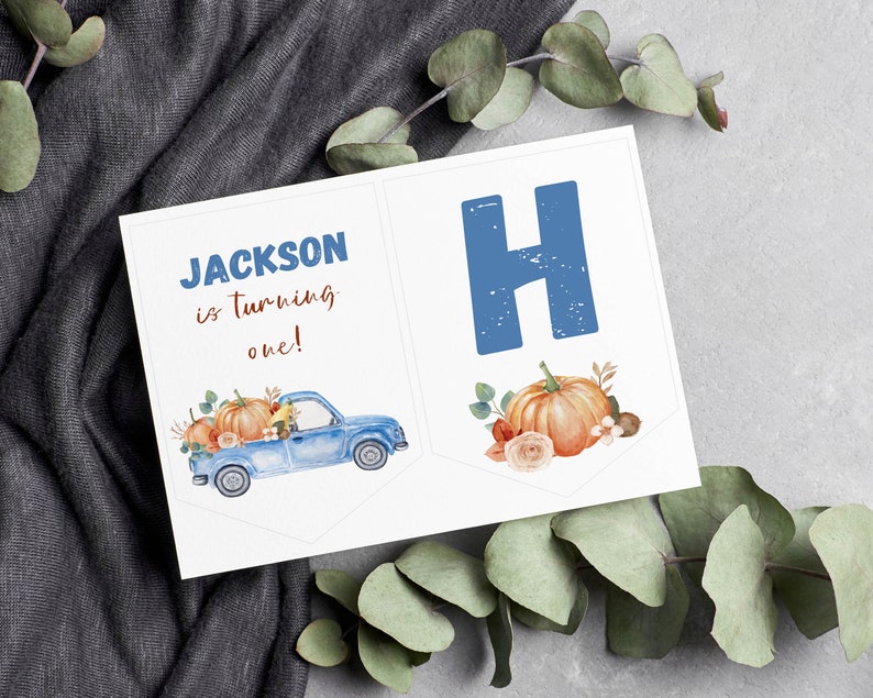 May include: A white rectangular banner featuring a blue truck carrying orange pumpkins and flowers, with the text "JACKSON is turning one!" in blue and brown, alongside a second banner section displaying a large blue letter H and an orange pumpkin.