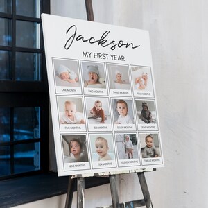 Op de afbeelding: Een wit rechthoekig fotobord met de zwarte tekst Jackson MY FIRST YEAR. Het bevat twaalf vierkante fotolijsten in drie rijen, elk gelabeld met een maand van &eacute;&eacute;n tot twaalf, met diverse portretten van zuigelingen in grijstinten.