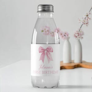 Puede incluir: Una botella de agua de pl&aacute;stico transparente con tapa de rosca plateada. La etiqueta presenta una ilustraci&oacute;n de un lazo en acuarela rosa sobre el texto Juliana's FIRST BIRTHDAY en una fuente serif rosa, centrado sobre un fondo blanco.