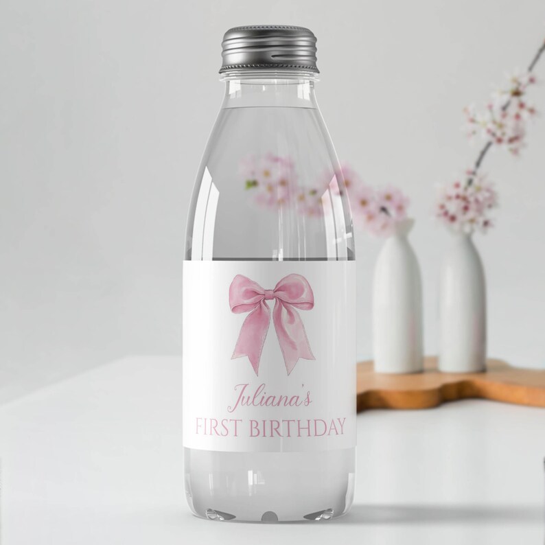 Puede incluir: Una botella de agua de pl&aacute;stico transparente con tapa de rosca plateada. La etiqueta presenta una ilustraci&oacute;n de un lazo en acuarela rosa sobre el texto Juliana's FIRST BIRTHDAY en una fuente serif rosa, centrado sobre un fondo blanco.