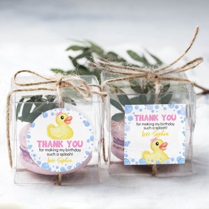 Pu&ograve; includere: Due scatole regalo quadrate trasparenti legate con spago, contenenti ciascuna un macaron viola chiaro e del verde. Le scatole presentano etichette con una papera di gomma gialla, bolle blu e il testo Thank you for making my birthday such a splash! Love Sophia.