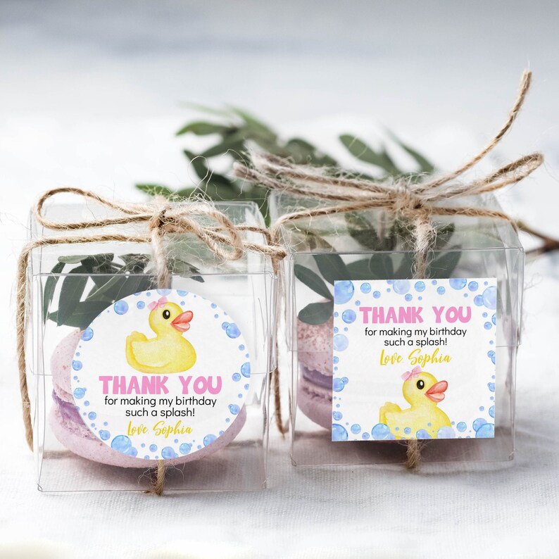 Pu&ograve; includere: Due scatole regalo quadrate trasparenti legate con spago, contenenti ciascuna un macaron viola chiaro e del verde. Le scatole presentano etichette con una papera di gomma gialla, bolle blu e il testo Thank you for making my birthday such a splash! Love Sophia.