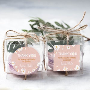 May include: Two clear square plastic favor boxes tied with brown twine, each containing a purple macaron and green leaves. The boxes feature pink labels with white daisy patterns and the text: THANK YOU For making my birthday so special! Love, Mila.