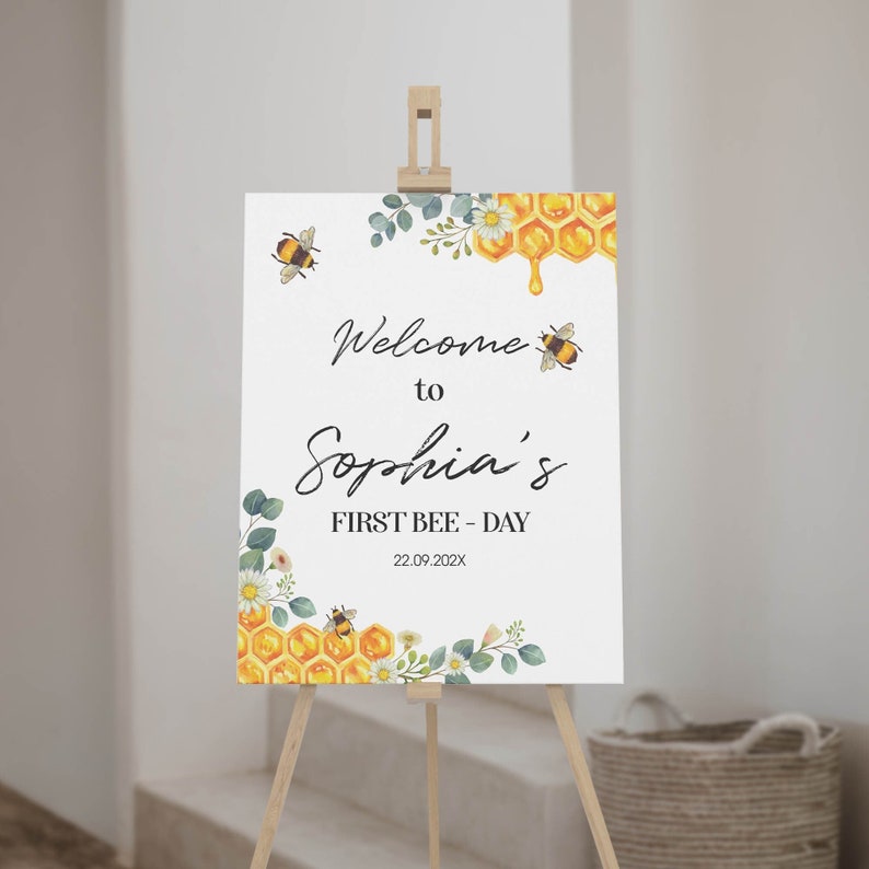 May include: A white sign on a wooden easel, featuring the text "Welcome to Sophia's FIRST BEE-DAY" and the date. The sign is decorated with watercolour bees, honeycomb, and floral accents.