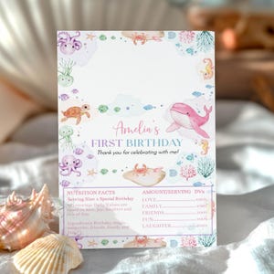 May include: A rectangular birthday card featuring watercolor sea creatures including a pink whale, purple octopus, and sea turtle. The text reads Amelia's FIRST BIRTHDAY Thank you for celebrating with me! and includes a Nutrition Facts label section.
