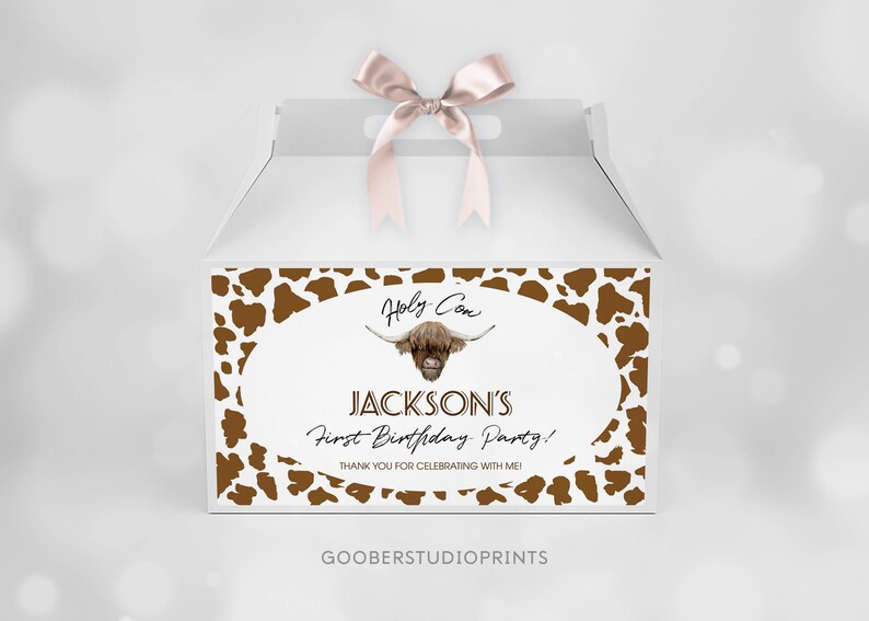 May include: A white gable box with a pink satin ribbon on top. The front features a brown cow print border surrounding an oval with a Highland cow illustration and the text: Holy Cow, JACKSON'S First Birthday Party! THANK YOU FOR CELEBRATING WITH ME!
