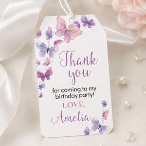 May include: A rectangular white gift tag with clipped corners and a white string, featuring watercolor-style butterflies in shades of purple and pink. The text reads: Thank you for coming to my birthday party! LOVE, Amelia, in purple script and black print.