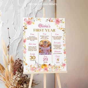 May include: A rectangular milestone sign titled Olivia's First Year, decorated with pink and white floral illustrations and pumpkins. It features text sections for interests, songs, foods, weight of 30 pounds, height of 29 inches, and 4 teeth.
