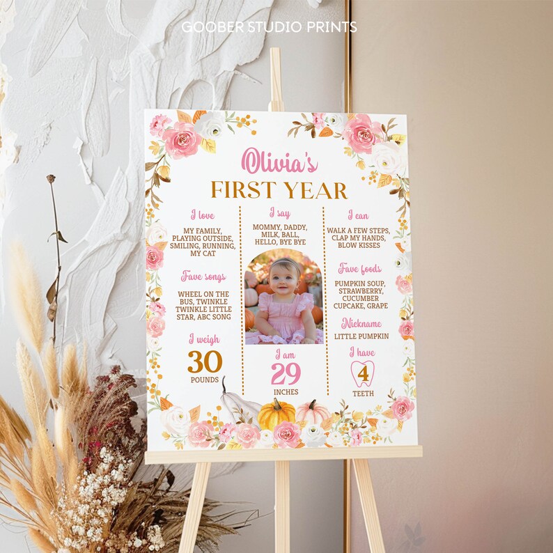 May include: A rectangular milestone sign titled Olivia's First Year, decorated with pink and white floral illustrations and pumpkins. It features text sections for interests, songs, foods, weight of 30 pounds, height of 29 inches, and 4 teeth.