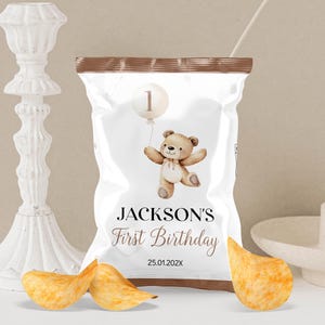 May include: A brown paper bag with a white label that says "JACKSON'S First Birthday" and has a teddy bear with a balloon that says "1" on it. The date "25.01.202X" is printed on the label.