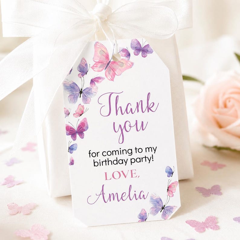 May include: A white rectangular gift tag with clipped corners featuring watercolor butterflies in shades of pink and purple. The text reads Thank you for coming to my birthday party! LOVE, Amelia. The tag is tied with a white ribbon.