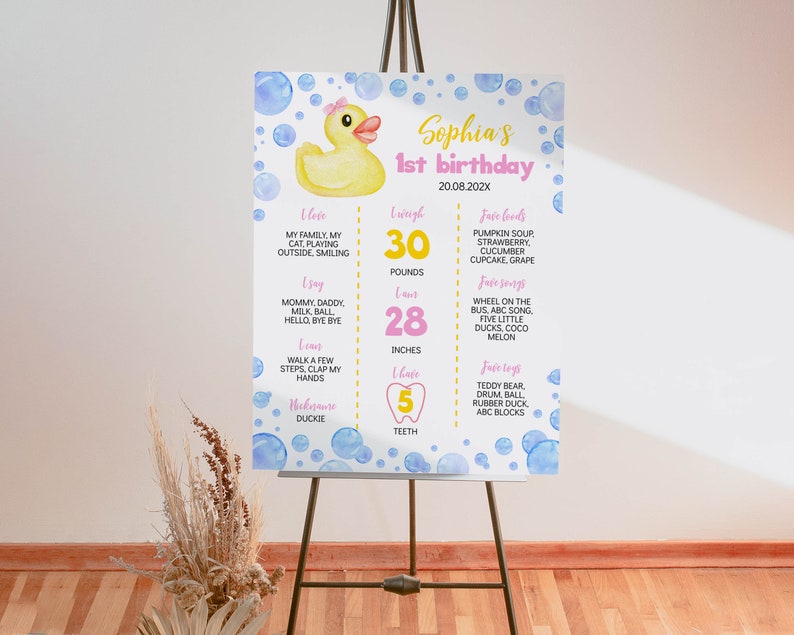 May include: A white birthday milestone sign featuring a yellow rubber duck with a pink bow, blue watercolor bubbles, and text including Sophia's 1st birthday, 30 pounds, 28 inches, and 5 teeth, displayed on a black metal easel stand.