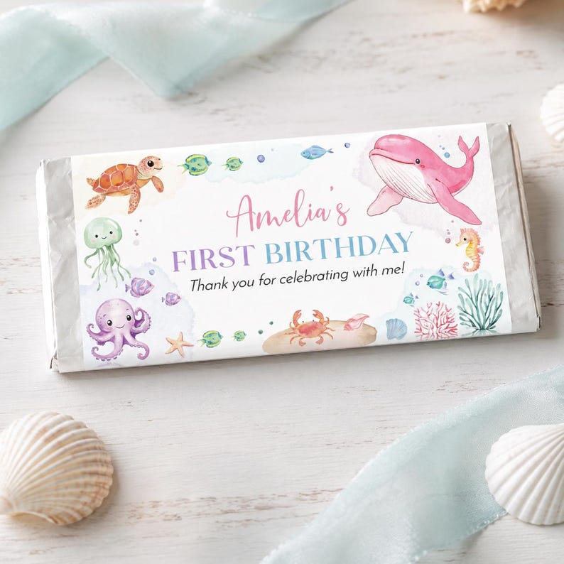 May include: A rectangular candy bar wrapper featuring watercolor illustrations of a sea turtle, jellyfish, octopus, crab, seahorse, and pink whale. The text reads Amelia's FIRST BIRTHDAY Thank you for celebrating with me! in pink and blue script.