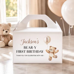 May include: A white cardboard gable box featuring a printed design of a brown teddy bear holding a balloon with the number 1. The text reads: Jackson's BEAR-Y FIRST BIRTHDAY THANK YOU FOR CELEBRATING WITH ME! The box has a top handle cutout.