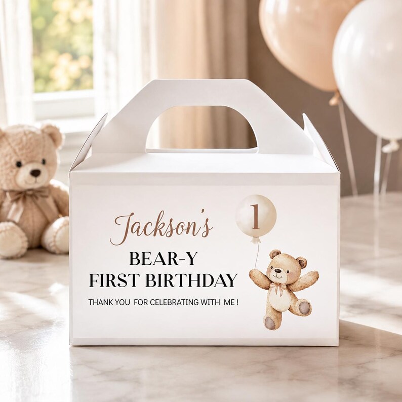 May include: A white cardboard gable box featuring a printed design of a brown teddy bear holding a balloon with the number 1. The text reads: Jackson's BEAR-Y FIRST BIRTHDAY THANK YOU FOR CELEBRATING WITH ME! The box has a top handle cutout.