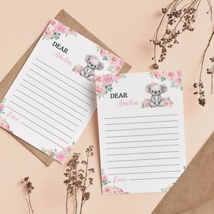 K&ouml;nnte beinhalten: Zwei wei&szlig;e Briefpapierb&ouml;gen mit horizontalen schwarzen Linien, verziert mit einer Aquarellillustration eines grauen Koalas mit rosa Blumenkranz, umgeben von rosa und pfirsichfarbenen Rosen und gr&uuml;nen Bl&auml;ttern. Text: DEAR Amelia und Love.