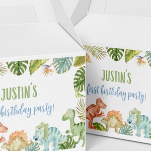 May include: Two white cardboard gable boxes featuring a watercolor jungle theme with green tropical leaves and colorful dinosaurs. The text on the boxes reads, "Justin's first birthday party!" in green and blue script and block lettering.