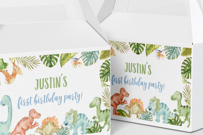 May include: Two white cardboard gable boxes featuring a watercolor jungle theme with green tropical leaves and colorful dinosaurs. The text on the boxes reads, "Justin's first birthday party!" in green and blue script and block lettering.