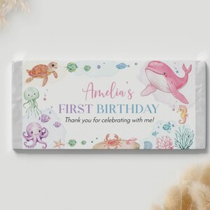 May include: A rectangular candy bar wrapper featuring watercolor illustrations of a pink whale, sea turtle, octopus, jellyfish, crab, and seahorse. The text reads Amelia's FIRST BIRTHDAY Thank you for celebrating with me! in pink and blue.