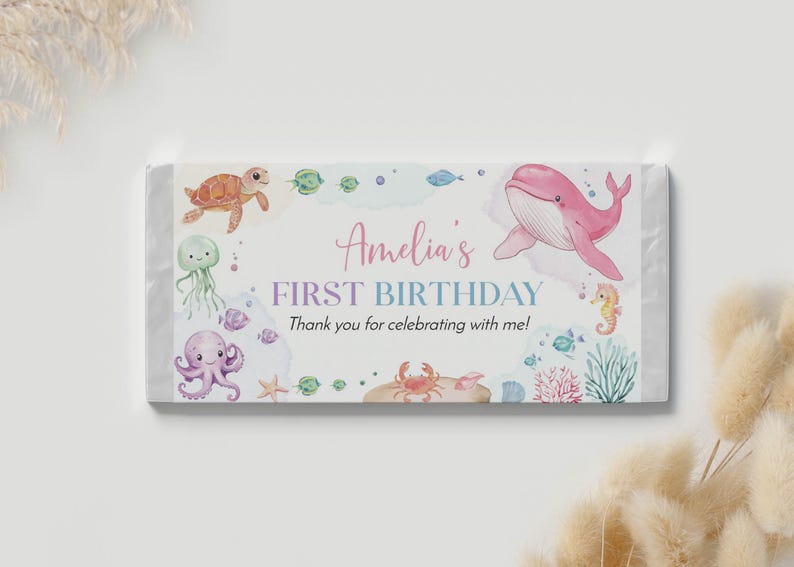 May include: A rectangular candy bar wrapper featuring watercolor illustrations of a pink whale, sea turtle, octopus, jellyfish, crab, and seahorse. The text reads Amelia's FIRST BIRTHDAY Thank you for celebrating with me! in pink and blue.
