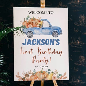 May include: A rectangular white sign featuring a blue pickup truck illustration carrying pumpkins and foliage. Text reads: WELCOME TO, one, JACKSON'S, First Birthday Party!, and 10.10.202X. The bottom border displays a decorative arrangement of pumpkins and leaves.