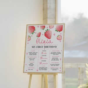 May include: A rectangular birthday milestone sign featuring pink strawberry illustrations and the name Olivia. The text includes My First Birthday, I Love, I Have 8 teeth, I Stand 35 inches, I Weigh 32 pounds, I Can, Fave Foods, Fave Songs, Fave Books, and Personality.