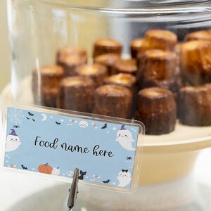 May include: A light blue rectangular food label card featuring a Halloween pattern with ghosts, pumpkins, bats, and spiderwebs, displaying the text Food name here, held by a metal clip in front of a glass dome filled with brown baked pastries.