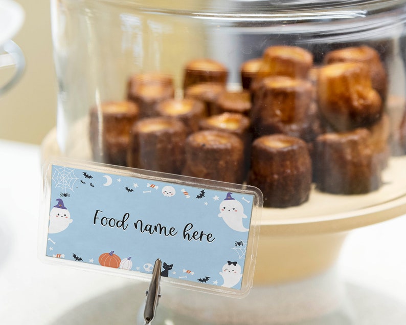 May include: A light blue rectangular food label card featuring a Halloween pattern with ghosts, pumpkins, bats, and spiderwebs, displaying the text Food name here, held by a metal clip in front of a glass dome filled with brown baked pastries.
