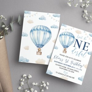 Pu&ograve; includere: Due biglietti d'invito di compleanno rettangolari con un'illustrazione ad acquerello di una mongolfiera blu e bianca che trasporta un orsacchiotto marrone. Il design include nuvole azzurre e beige, festoni e il testo Isnt he ONEderful.