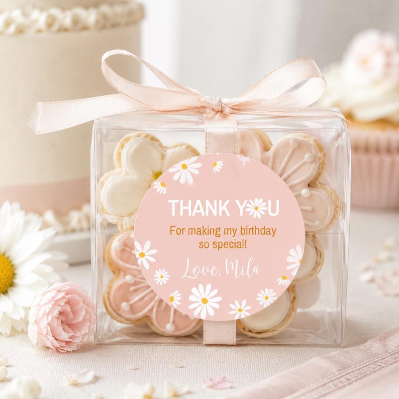 May include: A clear square gift box containing flower-shaped cookies, tied with a pale pink ribbon. A round sticker on the front features a pink background with white daisy illustrations and the text: THANK YOU For making my birthday so special! Love, Mila.