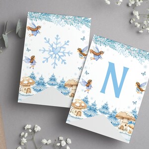 May include: Two white pennant flags featuring a winter theme with blue pine trees, mushroom houses, and flying fairies. One flag displays a large blue snowflake with a heart center, while the other features a large blue letter N in the center.
