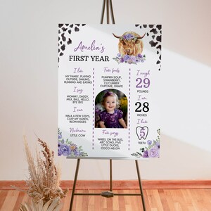 May include: A white milestone poster for Amelia's first year featuring a highland cow illustration with purple flowers, cow print corners, and text detailing favorite foods, songs, and milestones, including 29 pounds and 28 inches in black and purple.