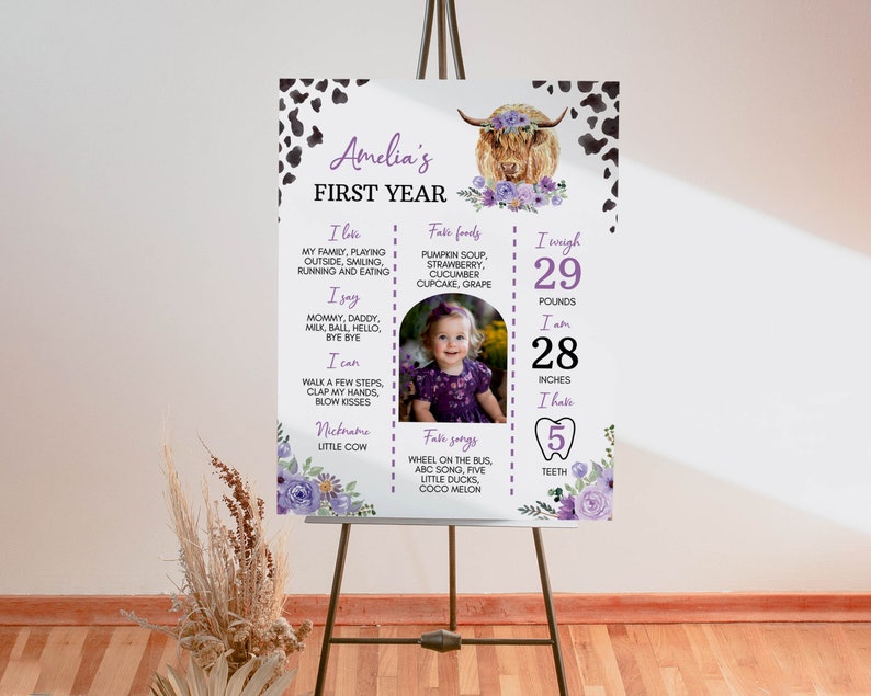 May include: A white milestone poster for Amelia's first year featuring a highland cow illustration with purple flowers, cow print corners, and text detailing favorite foods, songs, and milestones, including 29 pounds and 28 inches in black and purple.