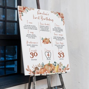 May include: A white milestone birthday sign for Amelia featuring orange pumpkin and floral illustrations. The text lists milestones including 30 pounds, 4 teeth, and 33 inches, alongside favorite foods, songs, and nicknames in a rust-colored font.
