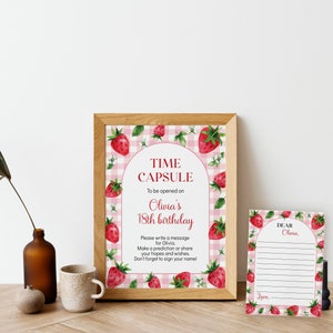 May include: A framed sign and a matching card featuring a pink gingham background with watercolor strawberry illustrations. The sign reads: TIME CAPSULE To be opened on Olivia's 18th birthday. The card reads: DEAR Olivia, with blank lines for writing.