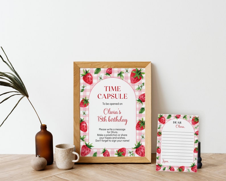 May include: A framed sign and a matching card featuring a pink gingham background with watercolor strawberry illustrations. The sign reads: TIME CAPSULE To be opened on Olivia's 18th birthday. The card reads: DEAR Olivia, with blank lines for writing.