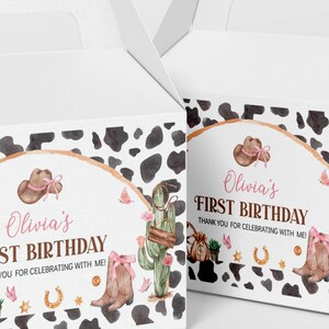 May include: Two white cardboard favor boxes featuring a cow print pattern and a watercolor design with a brown cowboy hat, pink bows, a cactus, a horseshoe, and a money bag. The text reads Olivia's FIRST BIRTHDAY THANK YOU FOR CELEBRATING WITH ME!