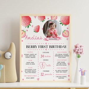 Op de afbeelding: Een gepersonaliseerde eerste verjaardag mijlpaalposter in een lichthouten lijst met aardbeienthema, een ronde foto van een kind en tekst met persoonlijke feiten zoals 34 inches, 5 tanden en 32 pounds, met roze en rode accenten.