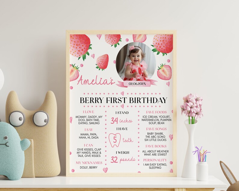 Op de afbeelding: Een gepersonaliseerde eerste verjaardag mijlpaalposter in een lichthouten lijst met aardbeienthema, een ronde foto van een kind en tekst met persoonlijke feiten zoals 34 inches, 5 tanden en 32 pounds, met roze en rode accenten.