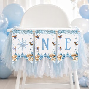 May include: A high chair banner featuring three white pennants with blue tulle trim. The design includes watercolor illustrations of fairies, blue snowflakes, and mushrooms. The letters O, N, and E are printed in large blue font across the pennants.