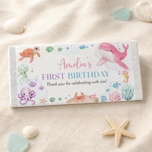 May include: A rectangular candy bar wrapper featuring watercolor illustrations of a pink whale, sea turtle, octopus, crab, and seahorse. The text reads Amelia's FIRST BIRTHDAY Thank you for celebrating with me! in pink and blue script and block fonts.