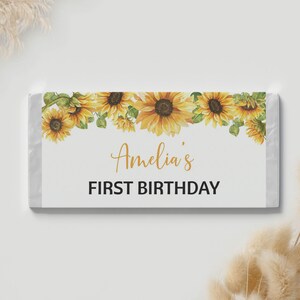 May include: A rectangular candy bar wrapper featuring a watercolor sunflower border along the top edge. The center of the white wrapper displays the text Amelia's in a gold script font, followed by the words FIRST BIRTHDAY in bold black text.