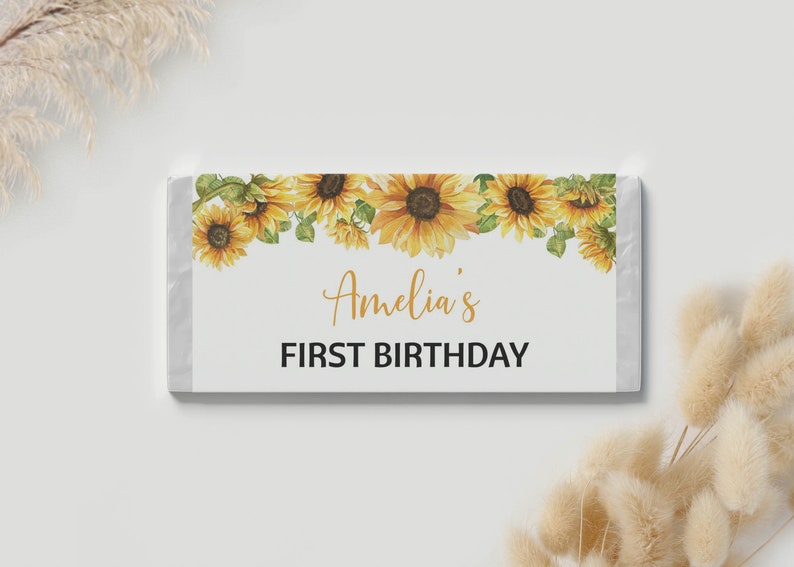 May include: A rectangular candy bar wrapper featuring a watercolor sunflower border along the top edge. The center of the white wrapper displays the text Amelia's in a gold script font, followed by the words FIRST BIRTHDAY in bold black text.