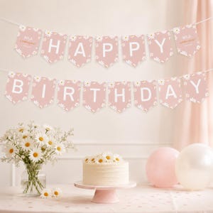 Op de afbeelding: Een tweedelige vlaggenlijn in oudroze met witte madeliefjespatronen en witte letters. De bovenste rij leest "MILA IS TURNING ONE" en de onderste rij "HAPPY BIRTHDAY". Elke vlaggetje heeft de vorm van een schild met een puntige onderkant.